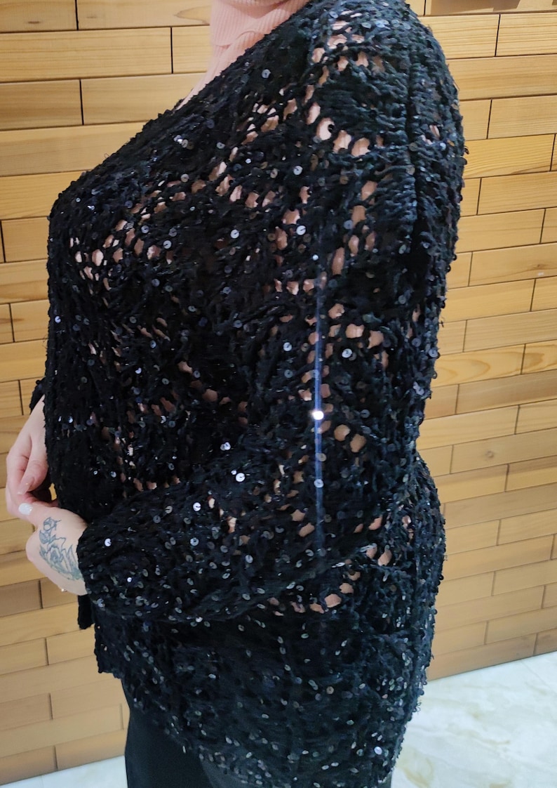 May include: A black, open-weave top with long sleeves, heavily embellished with sequins. The top has a loose fit. It is worn over a light-coloured garment. The background is a wooden wall.