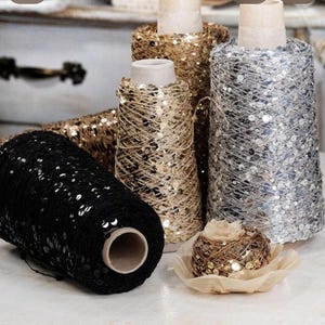 Könnte beinhalten: Eine Auswahl an Garnrollen mit Pailletten in verschiedenen Farben. Die Rollen sind in Schwarz, Gold und Silber gehalten. Das Garn ist um Pappkerne gewickelt, was einen strukturierten und schimmernden Effekt erzeugt. Ein kleines Deko-Objekt ist im Vordergrund.