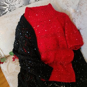 May include: A red, short-sleeved sweater with a high neck and sparkling sequins. A black, sequined shawl is draped over the sweater. The items are displayed on a patterned, cushioned surface.