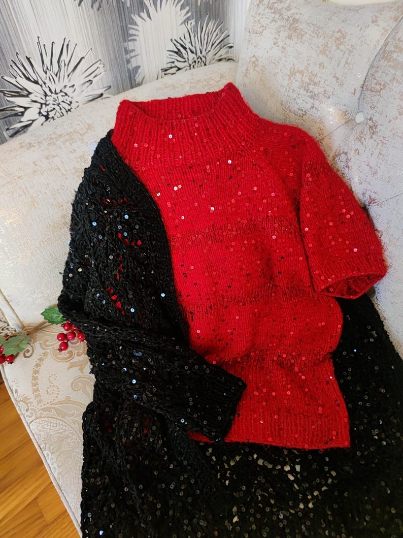 May include: A red, short-sleeved sweater with a high neck and sparkling sequins. A black, sequined shawl is draped over the sweater. The items are displayed on a patterned, cushioned surface.