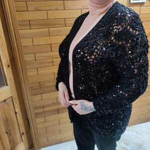 May include: Black open-front cardigan with sequin embellishments. The cardigan has a loose, open weave design and long sleeves. The garment is worn over a light pink turtleneck. The cardigan is a stylish addition to any wardrobe.