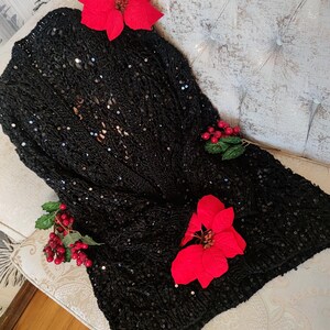 May include: A black sequined cardigan, open at the front, adorned with red poinsettia flowers and berry sprigs. The garment is displayed on a tufted, off-white upholstered surface, creating a festive aesthetic.