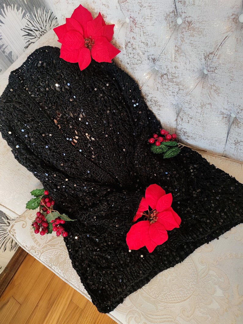 May include: A black sequined cardigan, open at the front, adorned with red poinsettia flowers and berry sprigs. The garment is displayed on a tufted, off-white upholstered surface, creating a festive aesthetic.