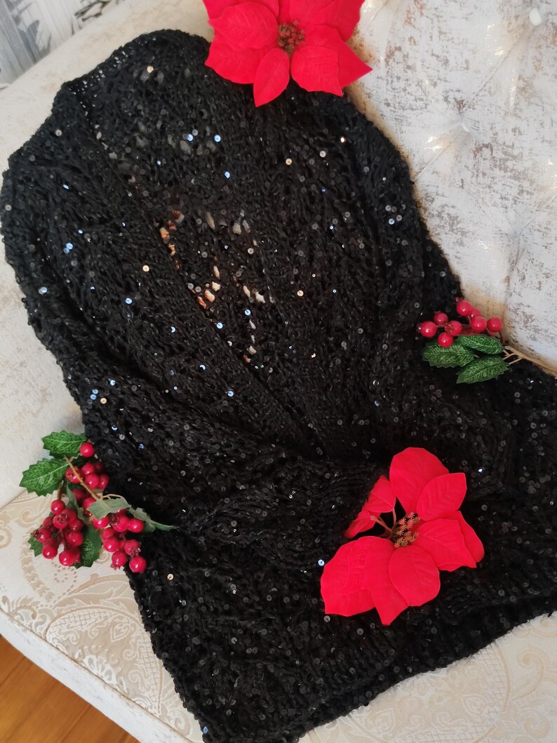 May include: A black sequined cardigan with an open front. The knitted garment is adorned with red poinsettias and berries, evoking a festive atmosphere. The sweater is presented on a patterned surface.