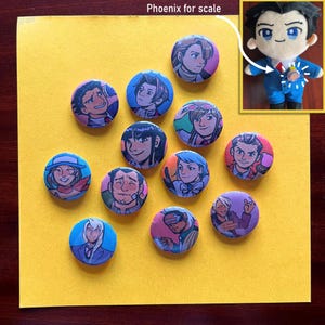 Può includere: Una collezione di bottoni rotondi e colorati con vari ritratti di personaggi in stile anime. I bottoni sono disposti su una superficie gialla, con un piccolo peluche di un personaggio nell'angolo in alto a destra, etichettato come "Phoenix for scale".