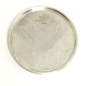 Może przedstawiać: Srebrny dekiel zegarka z wygrawerowanym tekstem, w tym "MADE IN SWITZERLAND" i nazwą marki. Okrągły element ma szczotkowane wykończenie i podniesioną krawędź na białym tle.