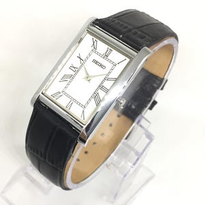 May include: A rectangular Seiko watch with a white face, black Roman numerals, and a silver-colored case. The watch has a black leather band with a crocodile pattern. The watch is displayed on a clear stand.