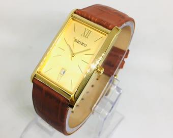 Reloj de pulsera Seiko Tank vintage: esfera dorada con ventana de fecha y correa de cuero color canela.