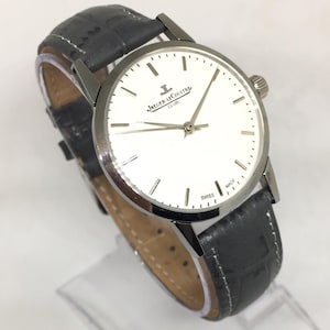 Może przedstawiać: Srebrny zegarek Jaeger-LeCoultre Club z białą tarczą i czarnym sk&oacute;rzanym paskiem. Zegarek ma okrągłą tarczę ze srebrnymi wskaz&oacute;wkami i znacznikami. Pasek jest teksturowany i ma zapięcie na sprzączkę. Zegarek jest oznaczony "SWISS MADE".
