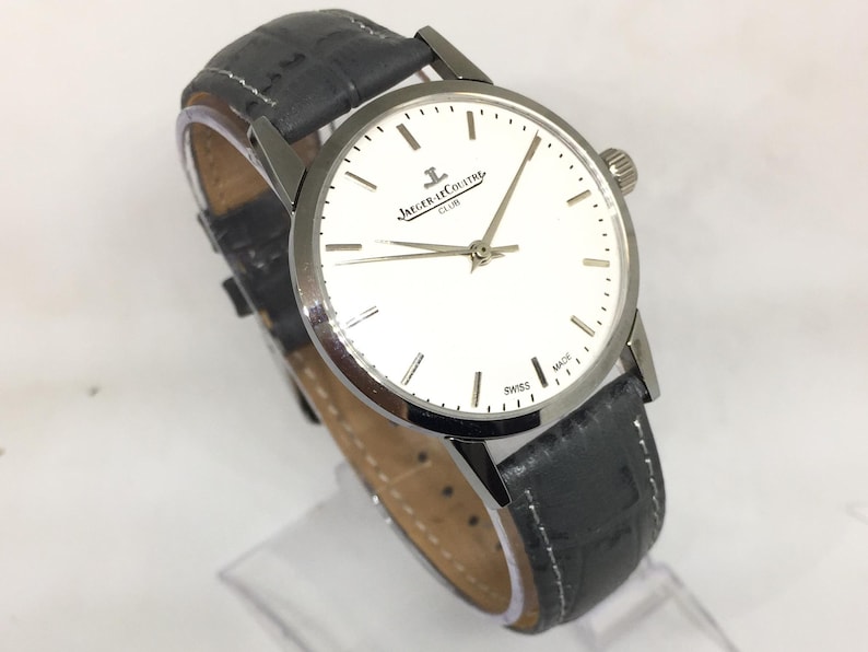 Może przedstawiać: Srebrny zegarek Jaeger-LeCoultre Club z białą tarczą i czarnym sk&oacute;rzanym paskiem. Zegarek ma okrągłą tarczę ze srebrnymi wskaz&oacute;wkami i znacznikami. Pasek jest teksturowany i ma zapięcie na sprzączkę. Zegarek jest oznaczony "SWISS MADE".