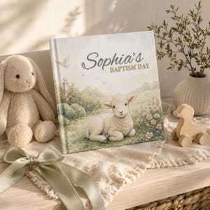 K&ouml;nnte beinhalten: Ein quadratisches Hardcover-Buch mit dem Titel Sophia's Baptism Day, das eine Aquarellillustration eines Lammes zeigt, das auf einer Wiese mit Blumen ruht. Das Cover enth&auml;lt eine kleine wei&szlig;e Taube sowie sanfte Gr&uuml;n- und Beiget&ouml;ne.