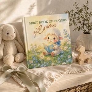 May include: A hardcover book titled First Book of Prayers for Emma, featuring an illustration of a lamb holding a blue book in a field of daisies and clover. The cover has a soft yellow and green color palette with a light blue bow on the lamb.