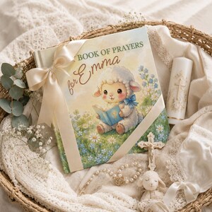 May include: A hardcover book titled MY BOOK OF PRAYERS for Emma featuring a cartoon lamb illustration, tied with a cream satin ribbon. Included are a white pillar candle with a gold cross, a silver crucifix, a pearl rosary, and a small plush lamb.