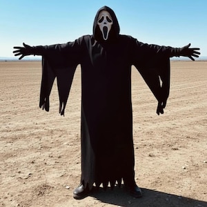 Puede incluir: Una persona con una túnica negra con capucha y una máscara de Scream se encuentra con los brazos extendidos en un paisaje desértico bajo un cielo azul claro. El disfraz incluye guantes y botas negras, creando un aspecto espeluznante para Halloween o cosplay.