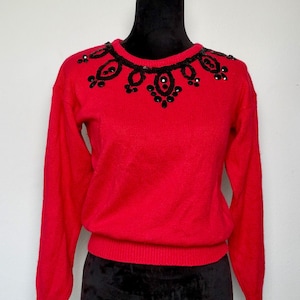 May include: A bright red sweater with long sleeves and a crew neck. The neckline is adorned with a black beaded design. The sweater is displayed on a black mannequin.