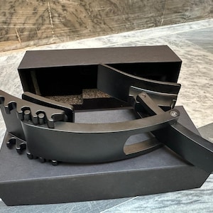 Könnte beinhalten: Eine mattschwarze Gürtelschnalle mit einzigartigem Design, die auf einer schwarzen Box liegt. Die Schnalle hat eine gebogene Form mit einer Reihe kleiner, abgerundeter Vorsprünge an einer Kante. Die Box ist geöffnet und zeigt ein dunkles Inneres.