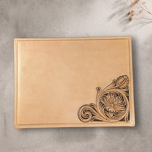 May include: A rectangular, light tan leather item with a decorative floral corner design. The design is dark brown and features a flower and swirling leaves. The edges are stitched with white thread.