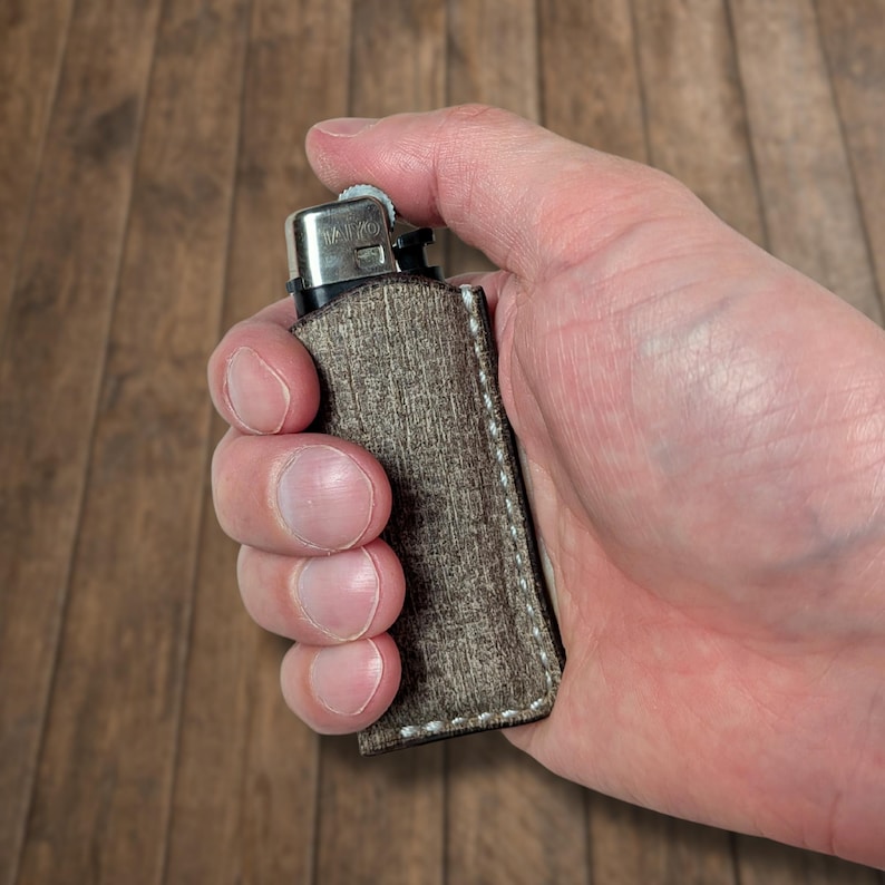 K&ouml;nnte beinhalten: Ein silberfarbenes Feuerzeug in einem strukturierten braunen Lederetui, das mit wei&szlig;em Faden gen&auml;ht ist. Das Feuerzeug wird in einer Hand vor einem Holzgrund gehalten.