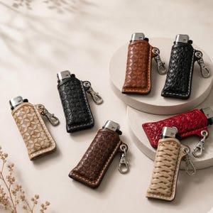 以下が含まれることがあります： 黒、茶色、ベージュなど、さまざまな色の革製ライターケースのコレクション。各ケースにはユニークなエンボス加工パターンと、銀色の金属クリップが付いています。ライターはニュートラルな背景に表示されています。