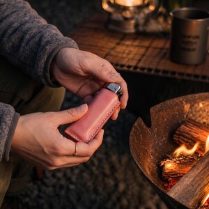 K&ouml;nnte beinhalten: Eine rosa Leder-Feuerzeugtasche mit wei&szlig;en N&auml;hten. Das Feuerzeug wird in den H&auml;nden einer Person in der N&auml;he eines Lagerfeuers gehalten. Die H&uuml;lle ist einfarbig und rechteckig. Das Feuerzeug ist silber und schwarz.