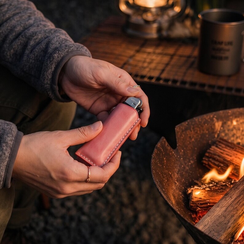 K&ouml;nnte beinhalten: Eine rosa Leder-Feuerzeugtasche mit wei&szlig;en N&auml;hten. Das Feuerzeug wird in den H&auml;nden einer Person in der N&auml;he eines Lagerfeuers gehalten. Die H&uuml;lle ist einfarbig und rechteckig. Das Feuerzeug ist silber und schwarz.