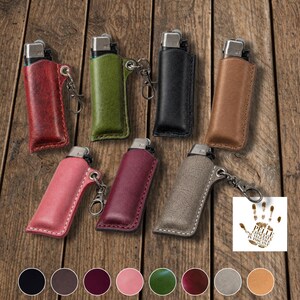 K&ouml;nnte beinhalten: Eine Auswahl an Leder-Feuerzeug-Etuis in verschiedenen Farben, darunter Rot, Gr&uuml;n, Schwarz und Braun. Jedes Etui hat einen silberfarbenen Metallclip. Die Feuerzeuge sind auf einer Holzoberfl&auml;che ausgestellt.