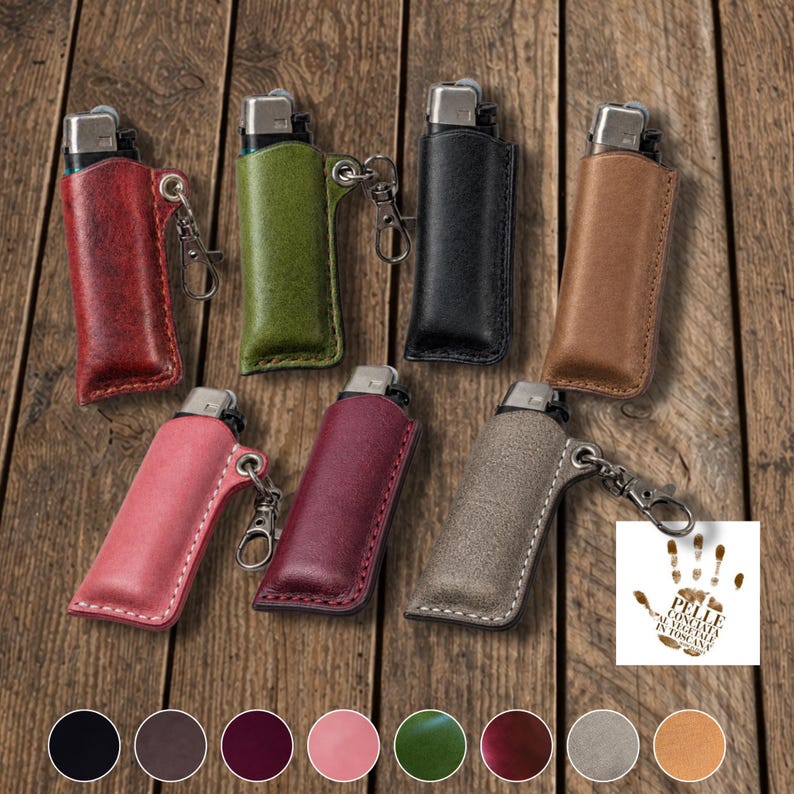 K&ouml;nnte beinhalten: Eine Auswahl an Leder-Feuerzeug-Etuis in verschiedenen Farben, darunter Rot, Gr&uuml;n, Schwarz und Braun. Jedes Etui hat einen silberfarbenen Metallclip. Die Feuerzeuge sind auf einer Holzoberfl&auml;che ausgestellt.