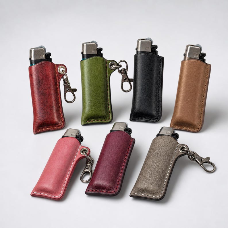 K&ouml;nnte beinhalten: Sieben Leder-Feuerzeug-Etuis in verschiedenen Farben, darunter Rot, Gr&uuml;n, Schwarz und Braun. Jedes Etui hat einen Metallverschluss und eine Schlaufe. Die Etuis sind auf wei&szlig;em Hintergrund angeordnet.