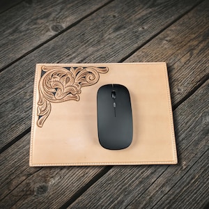 May include: A light tan leather mouse mat with a decorative floral design in the upper left corner and a black wireless mouse. The mouse mat has a stitched border and sits on a wooden surface.