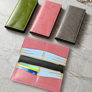 Op de afbeelding: Een verzameling leren portemonnees in verschillende kleuren, waaronder groen, roze en grijs. Eén portemonnee is open en toont kaartsleuven en een zwart interieur. De rechthoekige portemonnees hebben gestikte randen en een minimalistisch ontwerp.