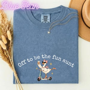 May include: A blue short-sleeved t-shirt featuring a graphic of a goose wearing sunglasses and a pink scarf, riding a scooter and holding a coffee cup. The text reads Off to be the fun aunt. A silver star necklace rests on the collar of the shirt.