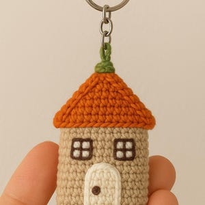May include: A handmade crochet house keyring. The house features an orange roof, beige walls, and a green detail. It has two windows and a door. The keyring is attached to a silver metal ring.
