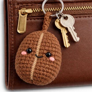 May include: A brown crocheted coffee bean keyring with a smiling face and pink cheeks. The keyring is attached to a silver ring with two keys. The background is a brown leather wallet.