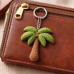 May include: A crochet palm tree keyring with a silver-coloured ring and chain. The palm tree has a brown trunk and green leaves. The keyring is set against a brown leather wallet with a gold zip. The wallet is a rich, warm colour.