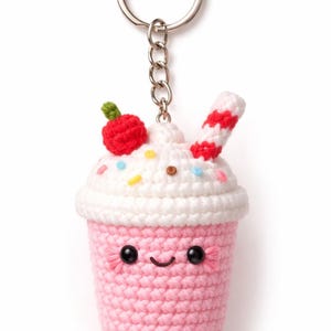 May include: A handmade crochet keychain shaped like a pink milkshake with a smiling face. The milkshake has white whipped cream, a red cherry, and a red and white striped straw. The keychain is attached to a silver metal ring and chain.