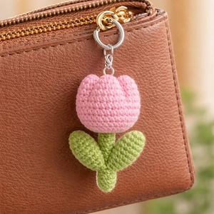 May include: A pink crochet tulip keychain with green leaves and stem, attached to a silver keyring. The keychain is displayed against a brown leather wallet with a gold zipper.