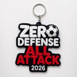 May include: A crocheted keyring featuring the text ZERO DEFENSE in white, ALL ATTACK in red, and the number 2026 in white, all outlined in black. A small crocheted football is attached to the top right, and a silver metal ring is at the top.
