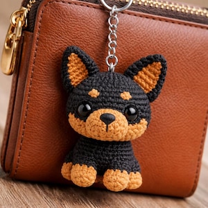 Puede incluir: Un llavero de perro de ganchillo negro y marrón con una cadena plateada. El perro hecho a mano tiene grandes ojos negros y está sentado frente a una cartera de cuero marrón. Las orejas, el hocico y las patas del perro son marrones.