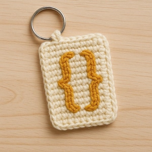 Könnte beinhalten: Gehäkelter Schlüsselanhänger mit cremefarbenem Hintergrund und einem senfgelben geschweiften Klammersymbol. Der Anhänger hat einen silbernen Metallring. Das handgefertigte Objekt liegt auf einer hellen Holzoberfläche.