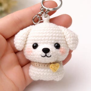 Puede incluir: Un llavero de perro blanco hecho a mano con ganchillo, con un llavero plateado. El perro tiene ojos negros, nariz negra y mejillas rosadas. Tiene un collar beige con un corazón amarillo. El llavero se sostiene en una mano.