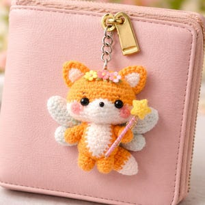May include: A crocheted fox keychain with a fairy theme. The fox is orange and white, with pink cheeks, a flower crown, and small wings. It holds a star wand. The keychain is attached to a pink wallet with a gold zipper.