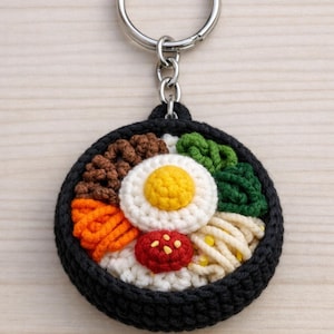 Puede incluir: Un llavero de ganchillo con un plato de bibimbap en miniatura. El plato incluye un huevo frito, verduras y arroz, todo en un cuenco negro. El llavero tiene un anillo plateado.