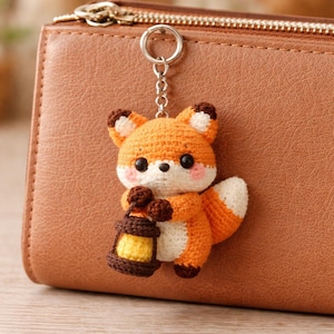 Könnte beinhalten: Ein gehäkelter Fuchs-Schlüsselanhänger in Orange, Weiß und Braun, der eine braune Laterne mit gelbem Schein hält. Der Fuchs hat schwarze Augen und rosa Wangen. Der Schlüsselanhänger ist an einem silbernen Ring befestigt, vor einem braunen Lederportemonnaie.