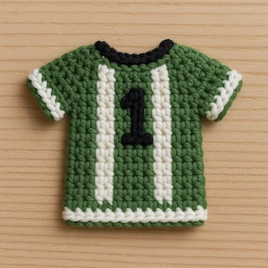 Puede incluir: Una camiseta de fútbol de ganchillo hecha a mano, verde y blanca, con el número 1 en negro. La camiseta tiene rayas y ribetes blancos, y un cuello negro. El artículo se muestra sobre una superficie de madera de color marrón claro.