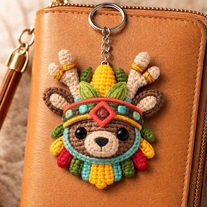May include: A handmade crochet keychain featuring a deer head with a colorful Native American headdress. The keychain has a brown, tan, yellow, red, and blue color scheme. The deer has large black eyes and is attached to a silver keyring.