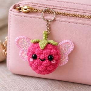 Può includere: Un portachiavi a forma di lampone fatto a mano all'uncinetto, con un viso sorridente, un gambo verde e ali rosa scintillanti. Il portachiavi rosa è attaccato a una borsa rosa con una cerniera dorata. Un accessorio carino.