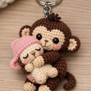 Puede incluir: Un llavero de mono de ganchillo hecho a mano en marrón, beige y rosa. El mono más grande abraza a un mono más pequeño que lleva un gorro rosa. El llavero tiene un anillo plateado adjunto a la parte superior de la cabeza del mono más grande.