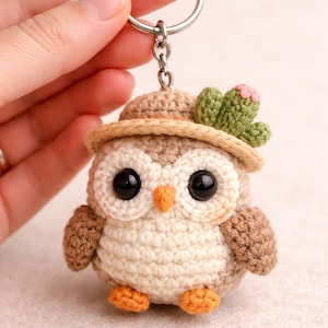 May include: A crocheted owl keychain with a small hat and cactus detail. The owl has large black eyes, an orange beak, and orange feet. The keychain is made of brown, beige, and green yarn.