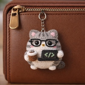 Puede incluir: Un llavero de gato tejido a ganchillo con gafas, sosteniendo una taza de café y un portátil que muestra símbolos de código. El gato es gris y blanco con detalles rosas, sujeto a un llavero plateado. El fondo es una cartera de cuero marrón.