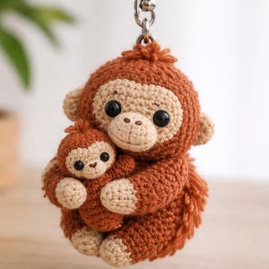 May include: A crochet keyring depicting a mother monkey embracing a baby monkey. The monkeys are brown with beige faces, hands, and feet, and have black eyes. The keyring is attached to a silver metal ring.